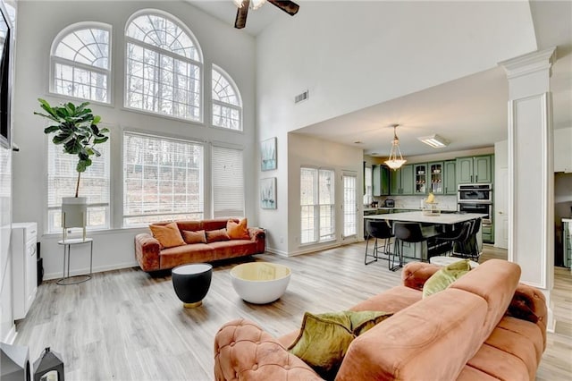 living room featuring baseboards, decorative columns, ceiling fan, light wood-style floors, and a towering ceiling