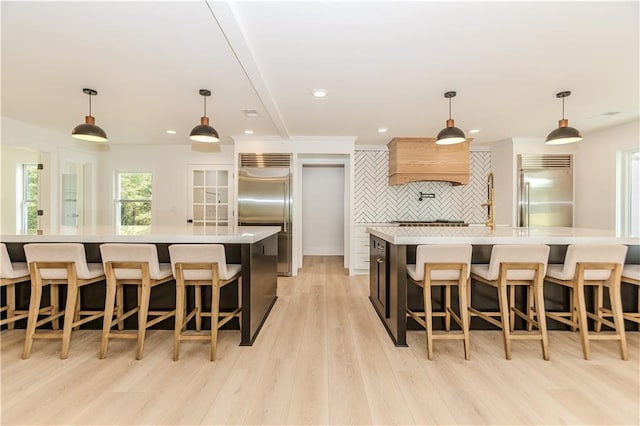 kitchen featuring hanging light fixtures, premium range hood, a large island with sink, decorative backsplash, and stainless steel built in fridge