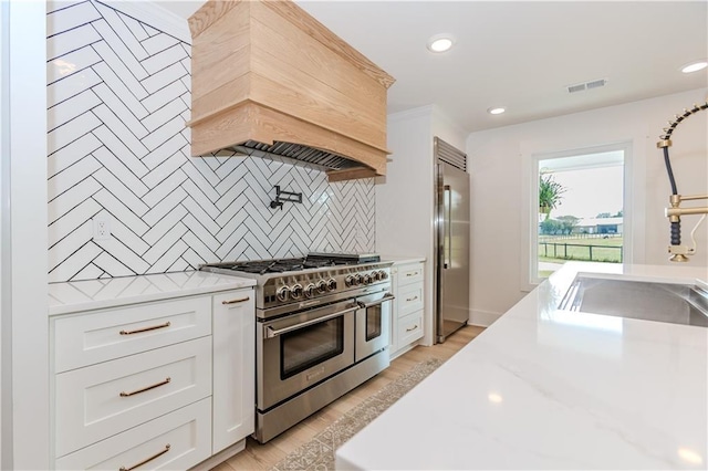 kitchen with custom exhaust hood, high end appliances, backsplash, white cabinets, and light stone countertops