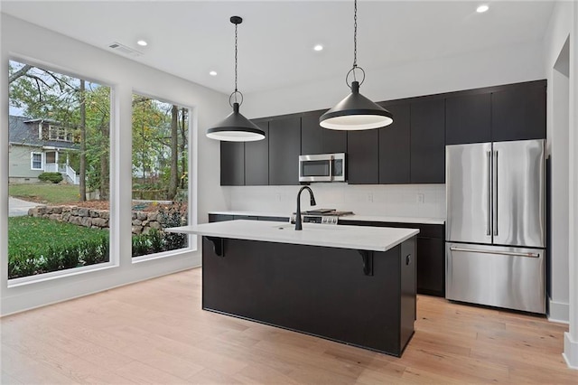 kitchen with a kitchen island with sink, light hardwood / wood-style flooring, pendant lighting, and appliances with stainless steel finishes