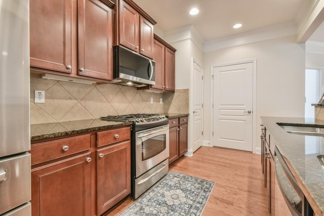 kitchen featuring stainless steel appliances, crown molding, dark stone countertops, light wood finished floors, and backsplash