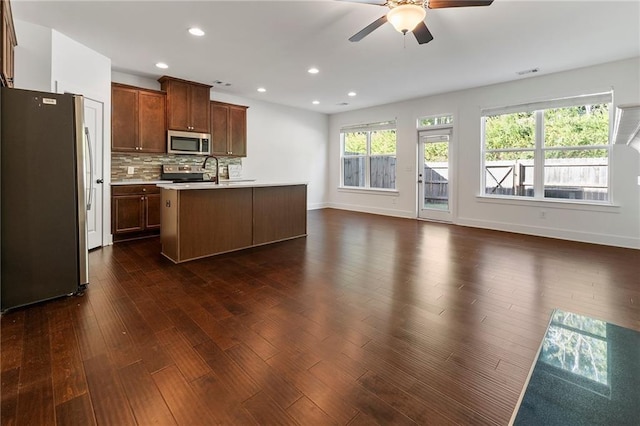 kitchen with ceiling fan, an island with sink, appliances with stainless steel finishes, tasteful backsplash, and dark hardwood / wood-style flooring