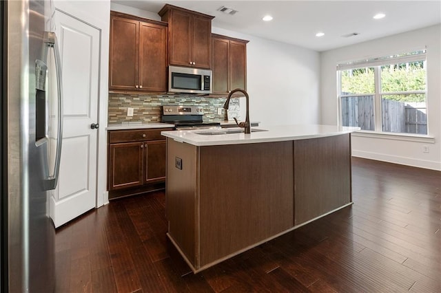 kitchen with appliances with stainless steel finishes, backsplash, a kitchen island with sink, sink, and dark hardwood / wood-style floors