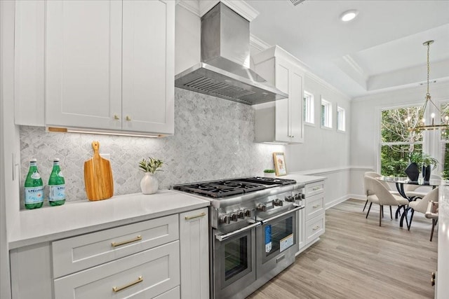 kitchen featuring wall chimney range hood, white cabinetry, double oven range, a tray ceiling, and decorative light fixtures