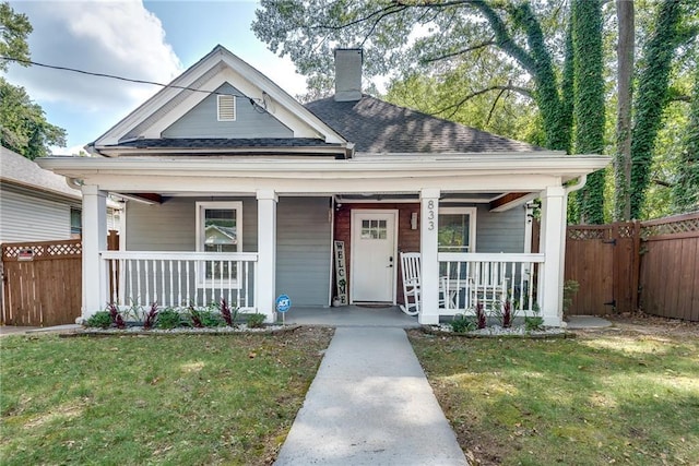 view of front of home with fence, roof with shingles, covered porch, a front yard, and a chimney