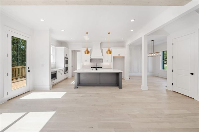 kitchen with hanging light fixtures, a kitchen island with sink, white cabinetry, and custom range hood