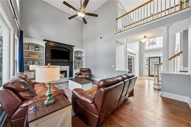 living room featuring ceiling fan, wood finished floors, baseboards, a tiled fireplace, and ornate columns