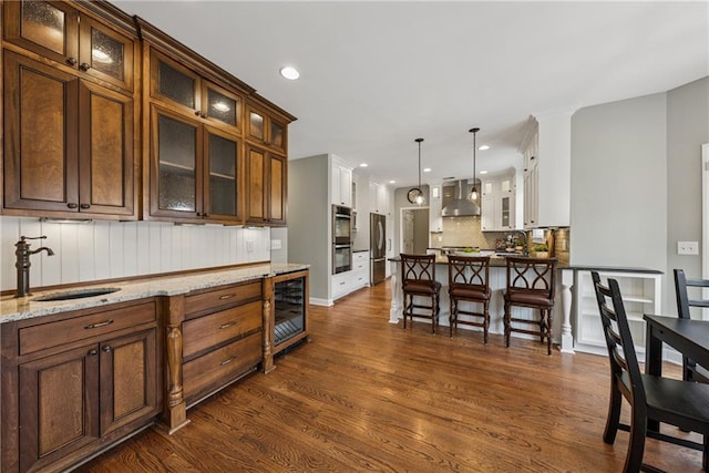 kitchen with beverage cooler, a sink, wall chimney range hood, appliances with stainless steel finishes, and dark wood finished floors