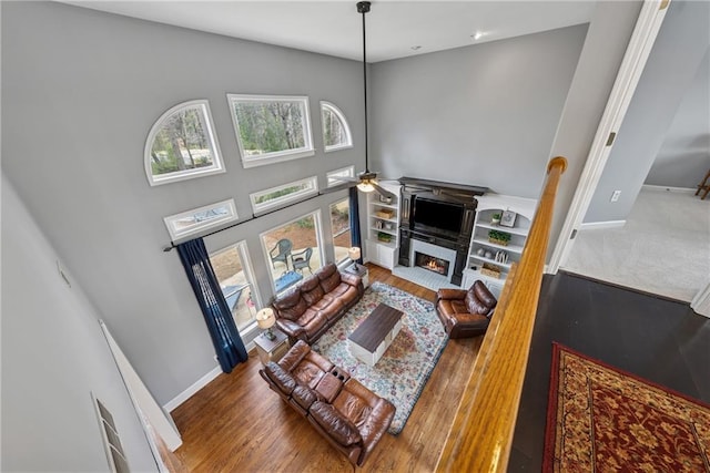 living area with ceiling fan, wood finished floors, a towering ceiling, baseboards, and a glass covered fireplace