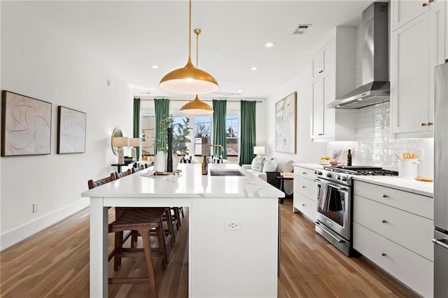 kitchen featuring high end range, wall chimney range hood, a center island with sink, white cabinetry, and hanging light fixtures