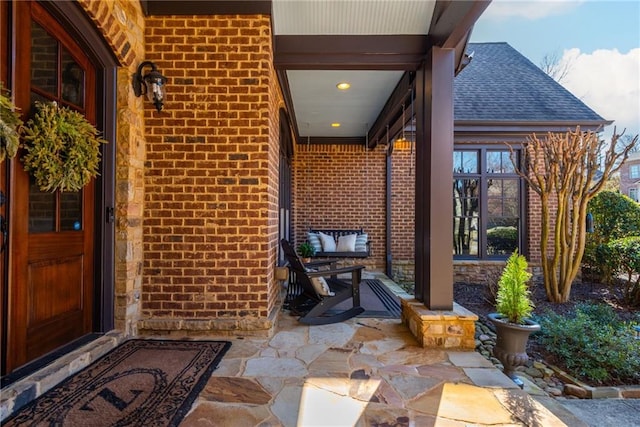 view of exterior entry featuring a porch, roof with shingles, and brick siding