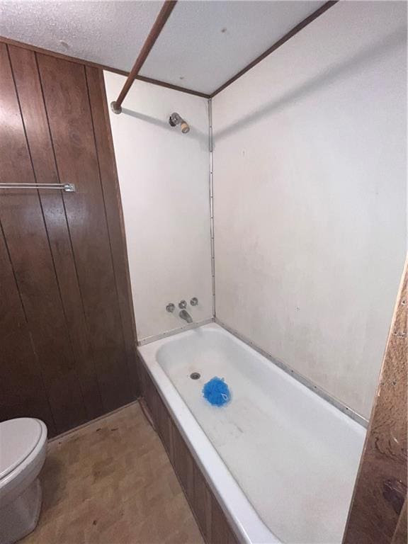 bathroom featuring a textured ceiling, wooden walls, toilet, and shower / bathing tub combination