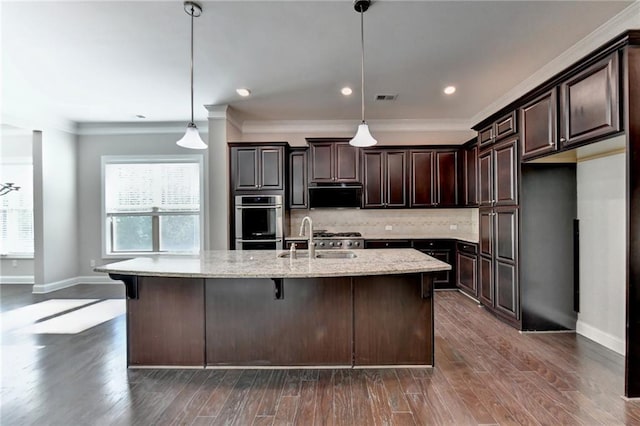 kitchen featuring pendant lighting, an island with sink, sink, dark brown cabinetry, and light stone counters