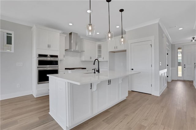 kitchen with hanging light fixtures, light countertops, wall chimney exhaust hood, an island with sink, and glass insert cabinets