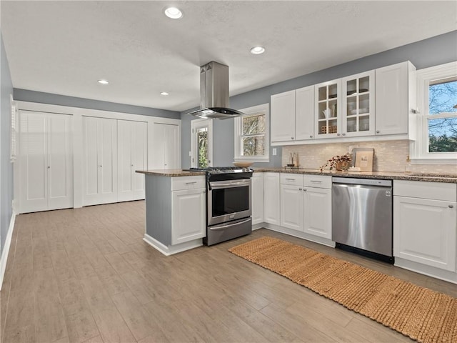 kitchen with a peninsula, glass insert cabinets, appliances with stainless steel finishes, light wood-type flooring, and island range hood