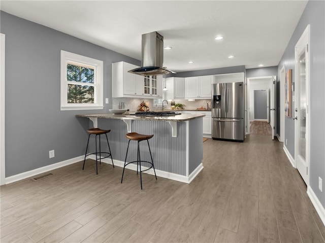 kitchen with tasteful backsplash, glass insert cabinets, a peninsula, stainless steel refrigerator with ice dispenser, and island range hood