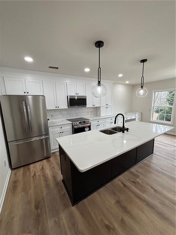 kitchen with sink, a kitchen island with sink, stainless steel appliances, white cabinets, and decorative light fixtures