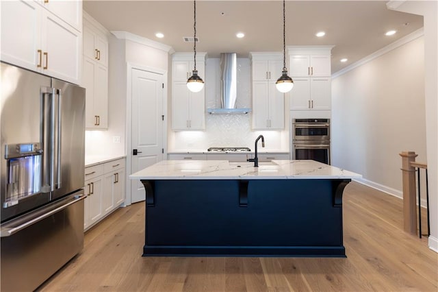 kitchen featuring appliances with stainless steel finishes, hanging light fixtures, light stone counters, a center island with sink, and wall chimney exhaust hood