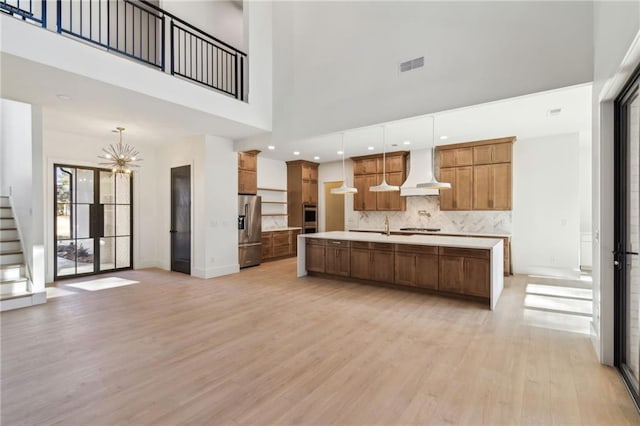 kitchen with wall chimney exhaust hood, a towering ceiling, decorative light fixtures, stainless steel appliances, and an island with sink