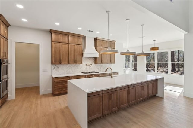 kitchen featuring stainless steel gas stovetop, premium range hood, light wood-type flooring, hanging light fixtures, and a large island with sink