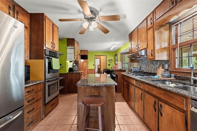 kitchen featuring under cabinet range hood, stainless steel appliances, a sink, a center island, and brown cabinets