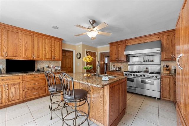 kitchen with stone counters, sink, wall chimney range hood, range with two ovens, and a center island with sink