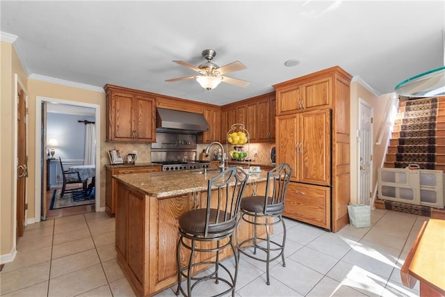 kitchen featuring ornamental molding, wall chimney exhaust hood, light tile patterned floors, stone counters, and an island with sink