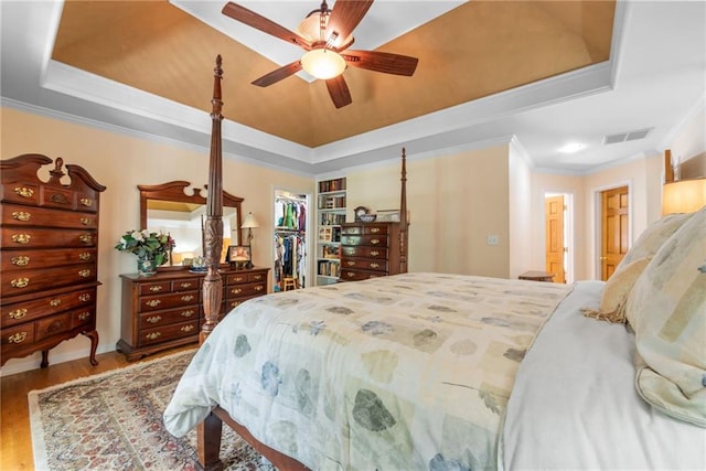 bedroom with a walk in closet, a raised ceiling, ceiling fan, crown molding, and hardwood / wood-style floors