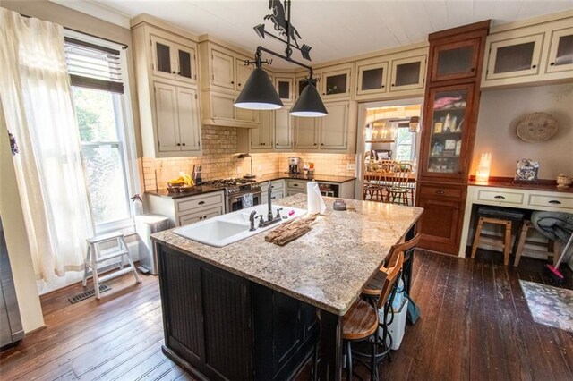 kitchen featuring hanging light fixtures, tasteful backsplash, a kitchen island with sink, cream cabinetry, and dark hardwood / wood-style floors