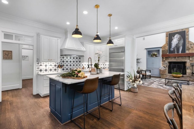 kitchen with stainless steel built in fridge, an island with sink, hanging light fixtures, white cabinetry, and dark hardwood / wood-style flooring