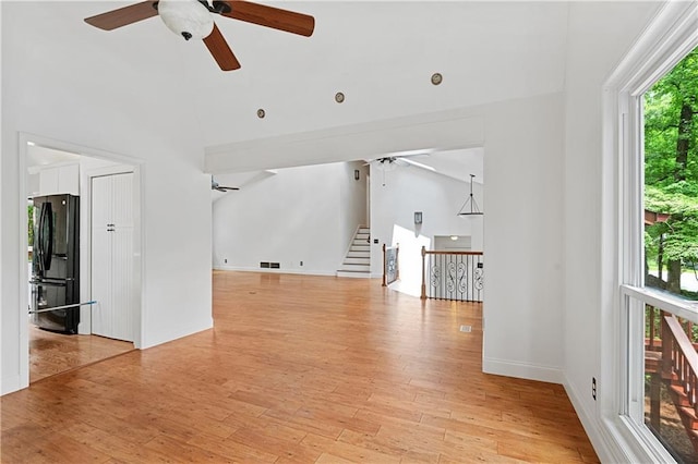 unfurnished living room with ceiling fan, light wood-style floors, baseboards, high vaulted ceiling, and stairway