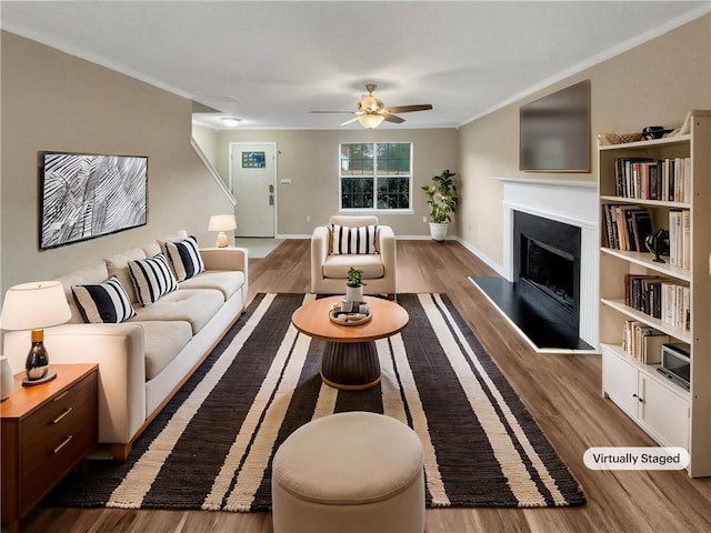 living area with wood finished floors, crown molding, a fireplace with raised hearth, and ceiling fan