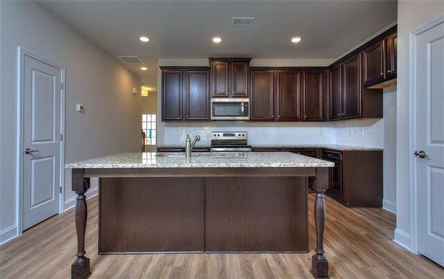 kitchen featuring light stone countertops, dark brown cabinets, a kitchen island with sink, stainless steel appliances, and light wood-type flooring