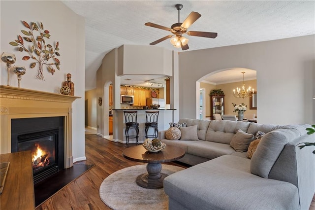 living area with dark wood finished floors, a glass covered fireplace, baseboards, arched walkways, and ceiling fan with notable chandelier