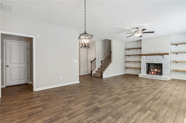 unfurnished living room featuring ceiling fan with notable chandelier, dark wood-type flooring, and a brick fireplace