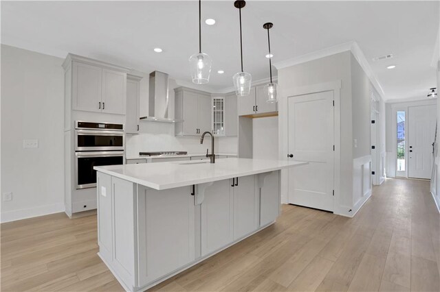 kitchen featuring hanging light fixtures, wall chimney range hood, a kitchen island with sink, stainless steel double oven, and sink