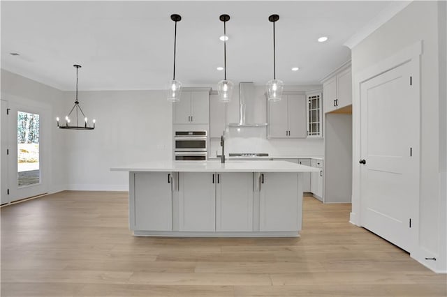 kitchen featuring double oven, pendant lighting, a center island with sink, and wall chimney exhaust hood