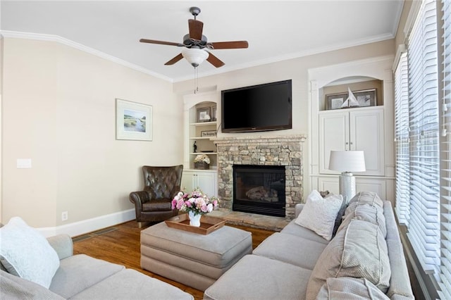 living room with built in shelves, ornamental molding, a stone fireplace, wood finished floors, and baseboards