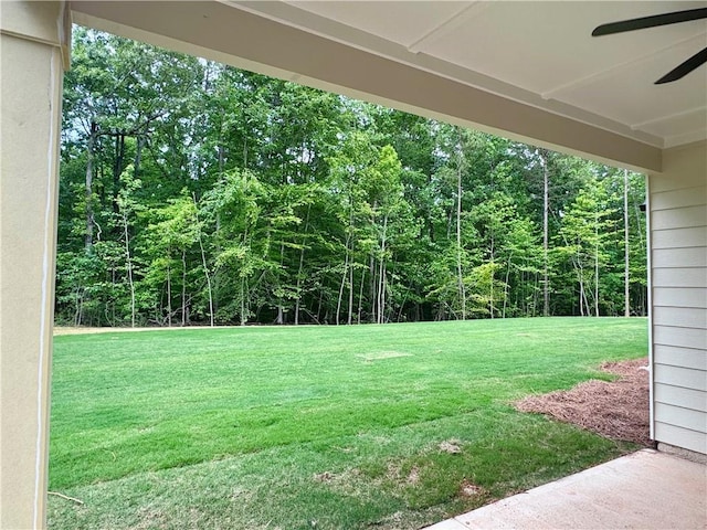 view of grassy yard featuring a ceiling fan and a wooded view