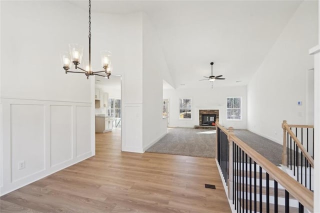 interior space featuring a decorative wall, a stone fireplace, high vaulted ceiling, light wood finished floors, and ceiling fan with notable chandelier