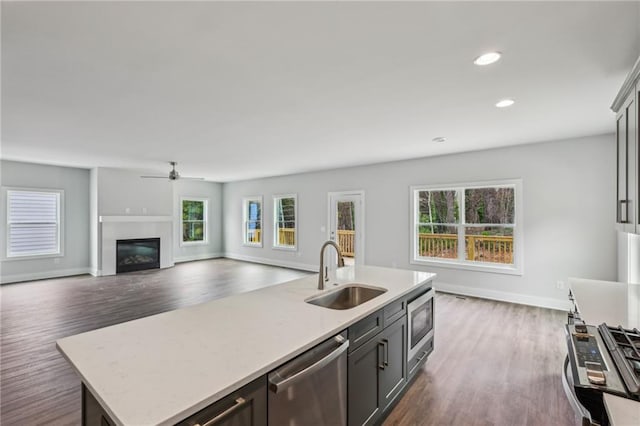 kitchen featuring an island with sink, a sink, a glass covered fireplace, stainless steel appliances, and dark wood-style flooring