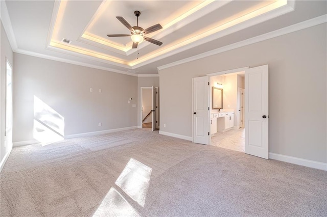 unfurnished bedroom featuring ceiling fan, a raised ceiling, crown molding, ensuite bathroom, and light carpet