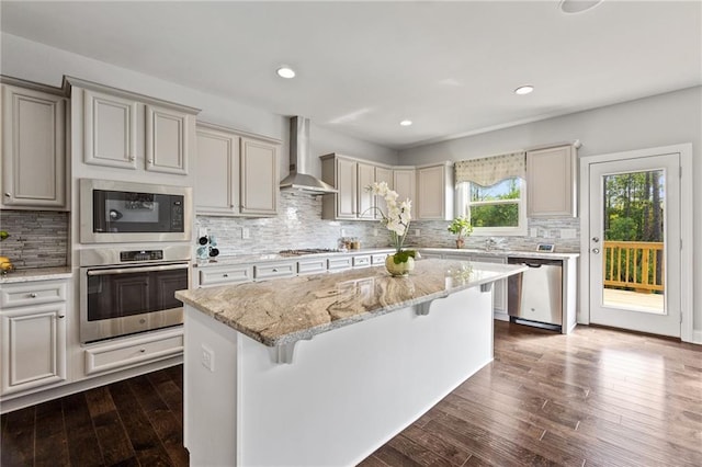 kitchen with wall chimney range hood, stainless steel appliances, a kitchen island, a kitchen bar, and backsplash
