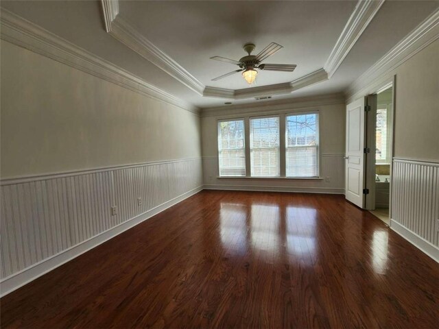 empty room with a ceiling fan, a wainscoted wall, wood finished floors, a tray ceiling, and crown molding