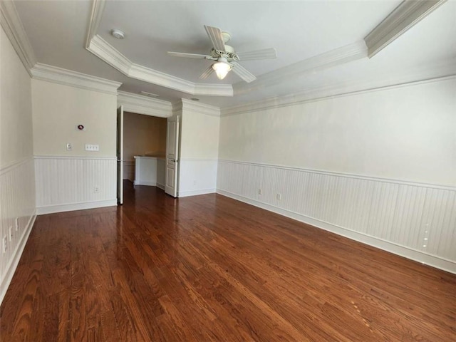 empty room featuring a tray ceiling, a wainscoted wall, a ceiling fan, ornamental molding, and wood finished floors