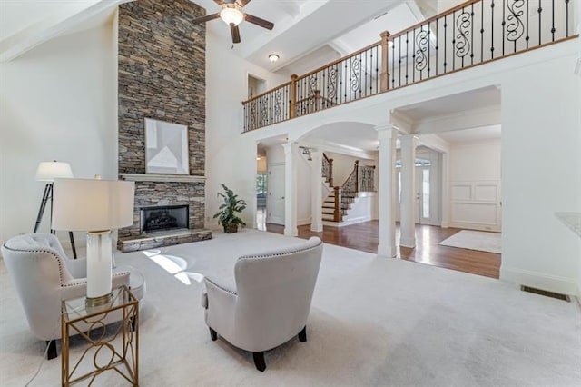 carpeted living room featuring a towering ceiling, decorative columns, ceiling fan, beam ceiling, and a stone fireplace