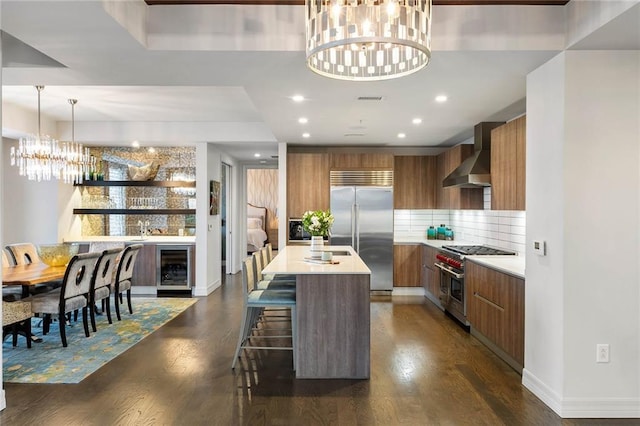 kitchen with wall chimney exhaust hood, dark wood-type flooring, premium appliances, an island with sink, and a chandelier