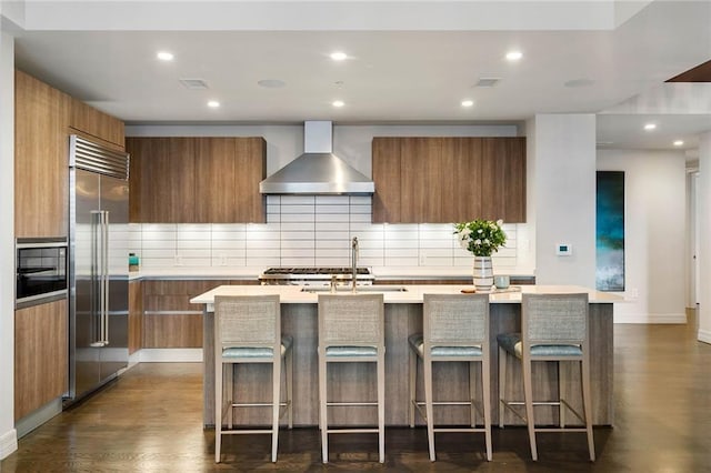 kitchen with a kitchen breakfast bar, wall chimney range hood, an island with sink, and built in refrigerator