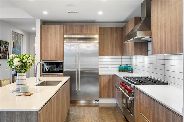 kitchen featuring backsplash, dark wood-type flooring, wall chimney range hood, sink, and built in appliances