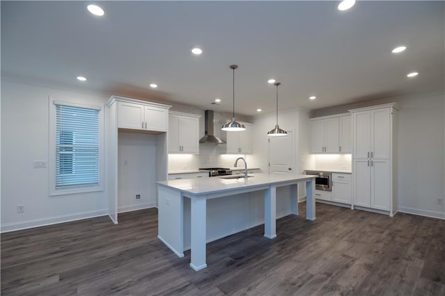 kitchen featuring pendant lighting, stainless steel appliances, wall chimney range hood, a kitchen island with sink, and white cabinetry